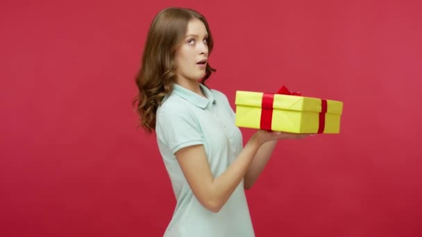 悪いプレゼント ポロシャツのブルネットの女性は慎重に 箱の中を見て 失望を表現し 驚きに不満をギフトをアンパック スタジオ ショットは赤の背景に隔離され — ストック動画