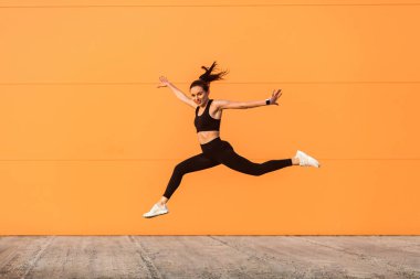Formda, spor kıyafetleriyle zıplıyor, koşuyor ve enerji dolu, sağlıklı spor yaşam tarzıyla gülümsüyor. Açık hava stüdyoları turuncu arka planda, reklam alanında izole edilmiş.