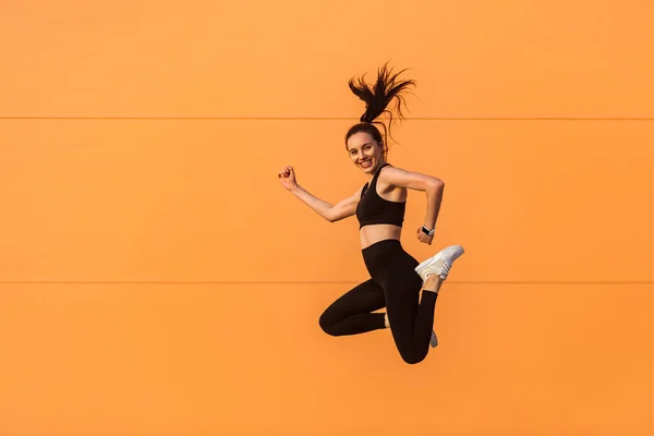 空気中でジャンプ 飛んで エネルギー 健康的なスポーツライフスタイルに満ちて笑みを浮かべてタイトなスポーツウェアでフィットボディと幸せな活気に満ちた鮮やかな女の子 スタジオ撮影 オレンジの背景に隔離された 広告エリア — ストック写真