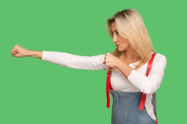 Tehditle mücadele et. Kendine güvenen, kot pantolon giymiş, boks tulumlu, kavgacı, saldırıyı püskürtmek için yumruklarını sıkan cesur sarışın kadın. Yeşil arka planda izole edilmiş kapalı stüdyo çekimi