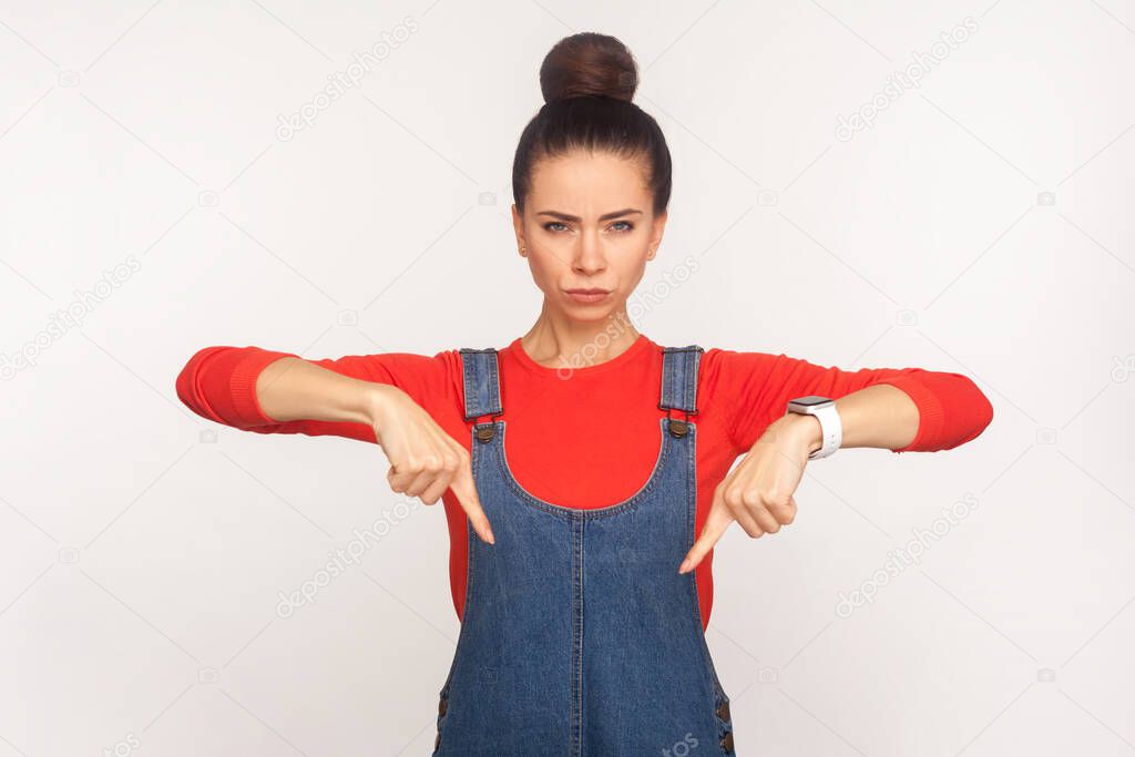 Do right now! Portrait of angry girl with hair bun in denim overalls pointing fingers down, demanding to come beside her immediately, giving order. indoor studio shot isolated on white background