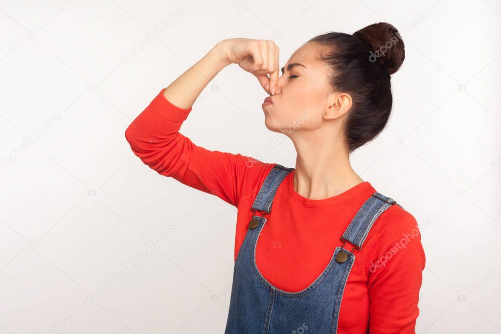 Repulsion to stinky odor. Portrait of girl with hair bun in denim overalls holding breath, pinching her nose and grimacing in disgust, avoiding bad smell. studio shot isolated on white background