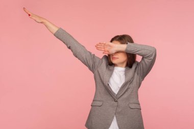 Portrait of successful young woman in business suit showing dab dance pose, famous internet meme of triumph, performing dabbing trends with hand gesture. studio shot isolated on pink background clipart