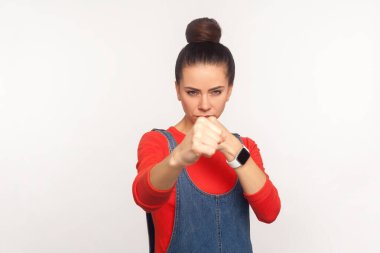Let's fight! Portrait of determined aggressive confident girl with hair bun in denim overalls boxing with clenched fist, punching in front of camera, struggle and self-defence. studio shot, isolate clipart