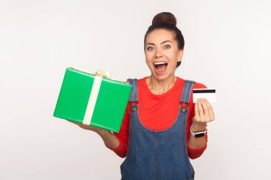 Wow, credit for holiday gift purchase! Portrait of stylish joyful girl with hair bun in denim overalls holding present box and bank card, enjoying shopping with cashback, bonus. studio shot isolated clipart