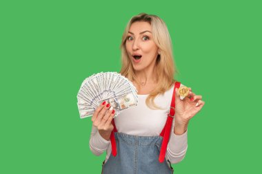 Kârlı dijital yatırım! Dolarlar ve btc sikke tutan şık tulumlu, bitcoin parasına karşı nakit para gösteren hayret verici yetişkin kadın portresi. Yeşil arka planda izole edilmiş kapalı stüdyo çekimi
