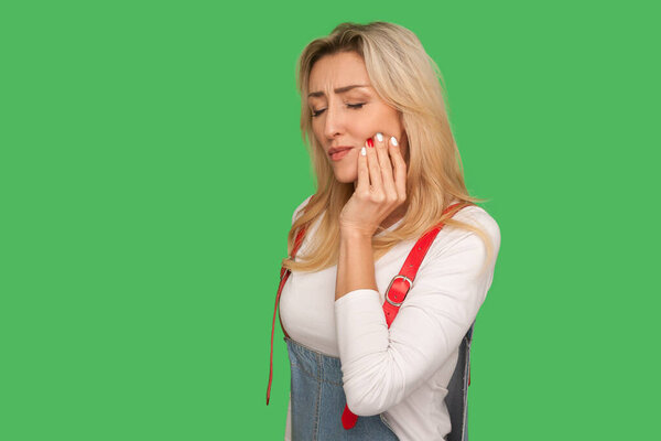 Dental problems, toothache. Portrait of sick adult woman in denim overalls touching sore cheek, suffering from cavities, cracked teeth, gum recession. indoor studio shot isolated on green background