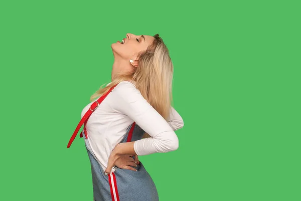 Spine problems. Portrait of unhealthy adult woman in denim overalls screaming in acute low back pain, suffering kidney inflammation, pinched nerve. indoor studio shot isolated on green background