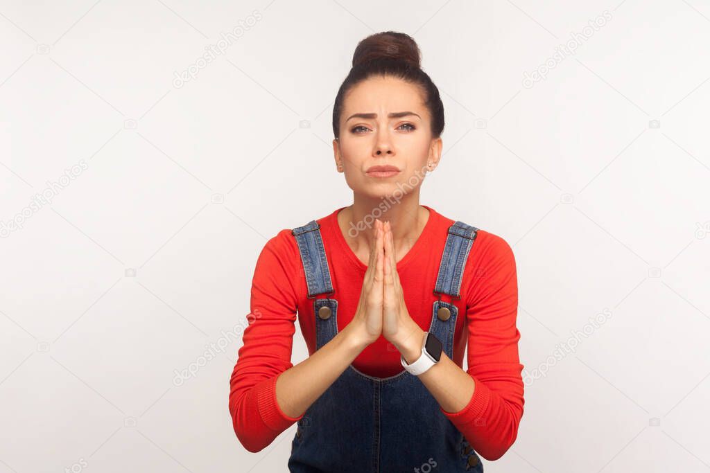 Please, I need! Portrait of imploring worried girl with hair bun in denim overalls begging, praying to camera with desperate pleading look, urging help. indoor studio shot isolated on white background