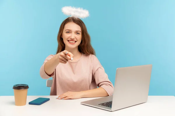 Lovely angelic positive woman employee with nimbus sitting at workplace, smiling and pointing to camera, indicating finger, choosing lucky winner. indoor studio shot isolated on blue background