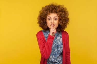 Sus, konuşma! Kıvırcık saçlı, dudakları parmaklarıyla şşşş yapan pozitif kadının portresi, sessiz kalmak istiyor, gizemli bir sır, konuşmak yasak. Sarı arka planda izole edilmiş kapalı stüdyo çekimi