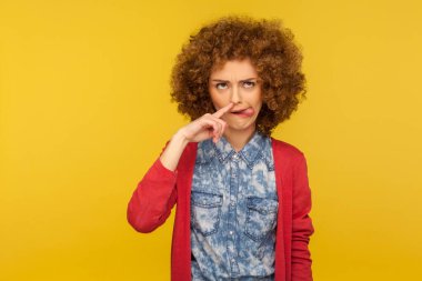 Kabarık kıvırcık saçlı, burnunu delen, dilini gösteren ve komik bir ifadeyle burun karıştıran komik kadının portresi. Sarı arka planda izole edilmiş kapalı stüdyo çekimi