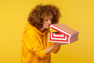 Portrait of upset displeased curly-haired woman in hoodie looking into gift box, opening present and peeking inside with dissatisfied expression. indoor studio shot isolated on yellow background clipart