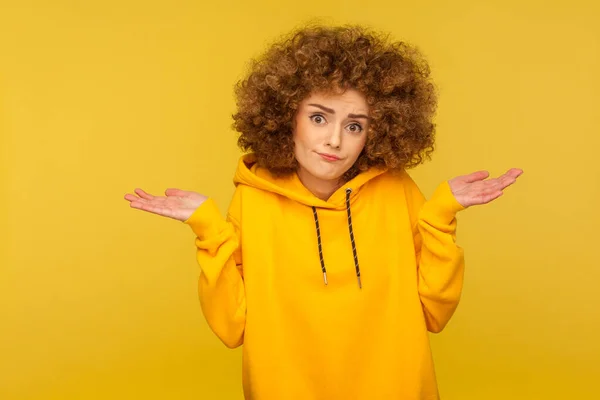 Don\'t know, sorry. Portrait of clueless uncertain confused curly-haired woman in urban style hoodie shrugging shoulders in questioning gesture, looking with indifference. indoor studio shot isolated