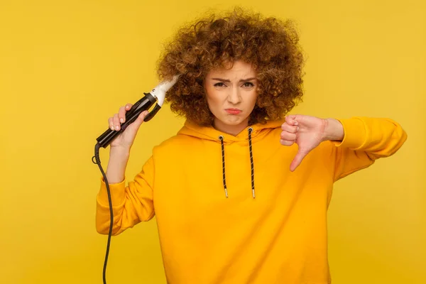 Upset displeased woman using curling iron to make afro hairdo and showing thumbs down dislike, ironing hair curls, expressing dissatisfaction with tool, bad hairstyling. indoor studio shot isolated