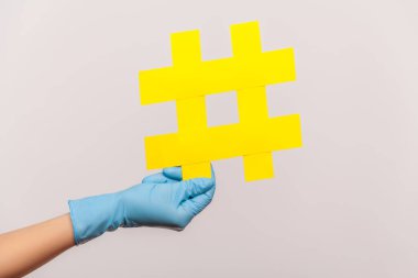 Profile side view closeup of human hand in blue surgical gloves holding yellow hashtag. indoor, studio shot, isolated on gray background. clipart