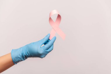 Profil yan görüntüsü, göğüs kanserinin sembolü olan mavi cerrahi eldivenli insan eli shwoing ve pembe kurdele tutuyor. İçerde, stüdyoda, gri arka planda izole edilmiş..