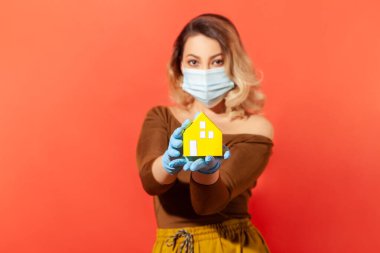 Koruyucu maskeli ve eldivenli bir kadın elinde kağıt ev, evde kalma tavsiyesi bulaşıcı hastalıklar yayılırken karantinaya alınma, koronavirüs salgını. Stüdyoda çekim izole edildi