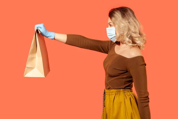 隔離された安全な買い物 紙袋を与える保護マスクのサイドビューの女性は 食品を購入するコロナウイルスの自己分離 オンライン注文で配信 赤の背景に隔離された屋内スタジオで — ストック写真