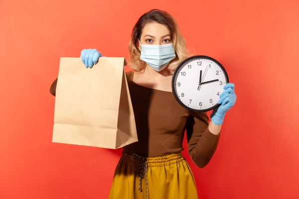 Time to order food, safe shopping on quarantine. Woman in protective mask holding paper bag and big clock, delivery in coronavirus self-isolation. indoor studio shoot isolated on red background