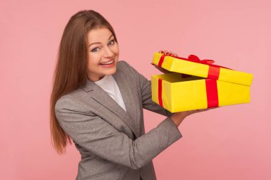 Portrait of attractive happy businesswoman in suit jacket opening gift box and looking excitedly in anticipation of surprise, unwrapping birthday present. studio shot isolated on pink background clipart