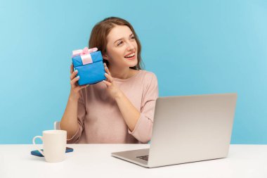 Pretty woman employee sitting at workplace and holding gift near ear, listening with interest what's inside box, curious about birthday surprise, bonus for good job. indoor studio shot, isolated clipart