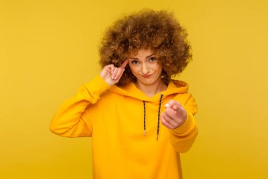 You are crazy! Portrait of curly-haired woman in urban style hoodie making stupid sign with finger near head and pointing to camera, gesturing bad mind, dumb insane idea. indoor studio shot, isolated
