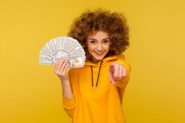 Start making money! Portrait of joyful curly-haired woman in urban style hoodie holding bunch of dollar banknotes and pointing to camera, encouraging to earn profit. indoor studio shot, isolated clipart