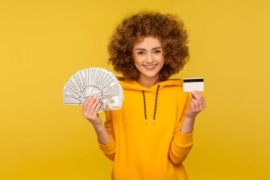 Para tasarrufu, banka hesabı. Şehir tarzı kapüşonlu mutlu kıvırcık saçlı kadın portresi dolar banknotları ve plastik banka kartı gösteriyor, kameraya gülümsüyor. Stüdyo çekimi sarı arkaplanda izole edildi