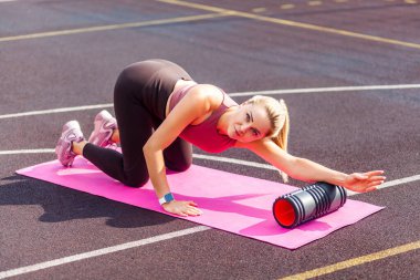 Sporcu, sarışın, sporcu bir kadın yaz günü paspas üzerinde antrenman yapıyor. Sırt ağrısı için köpük silindirli masaj aleti kullanıyor. El ve omurga kaslarını esnetiyor, fasya egzersizi yapıyor. Sağlık hizmetleri, egzersiz rutinleri
