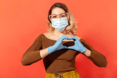 Hijyenik maske takan dost canlısı bir kadın, viral hastalığı önlemek için eldiven ve gözlük takıyor, kalp hareketi gösteriyor, Coronavirus 2019 ncov hastalarına sevgi desteği veriyor. Stüdyoda çekim izole edildi