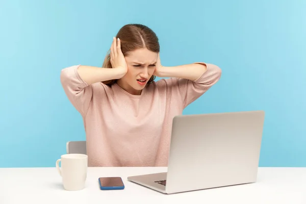 Don\'t want to hear you. Angry woman employee covering ears and looking irritably at laptop screen, annoyed by unpleasant communication on video call. indoor studio shot isolated on blue background