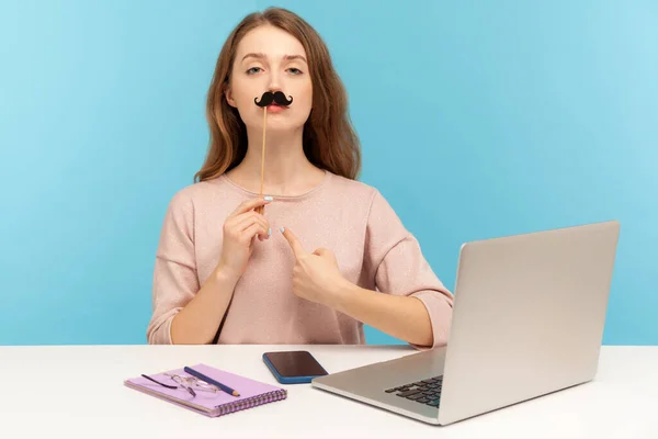Funny arrogant woman office employee pointing herself, covering lips with fake paper moustache, imitating strict boss, humorous masquerade accessory. indoor studio shot isolated on blue background