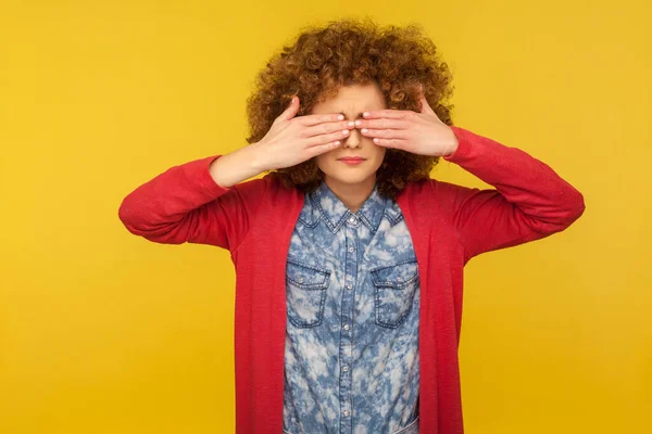 Don\'t want to look. Portrait of scared woman with curly hair in casual outfit covering eyes, feeling shy and afraid to watch, ignoring troubles. indoor studio shot isolated on yellow background
