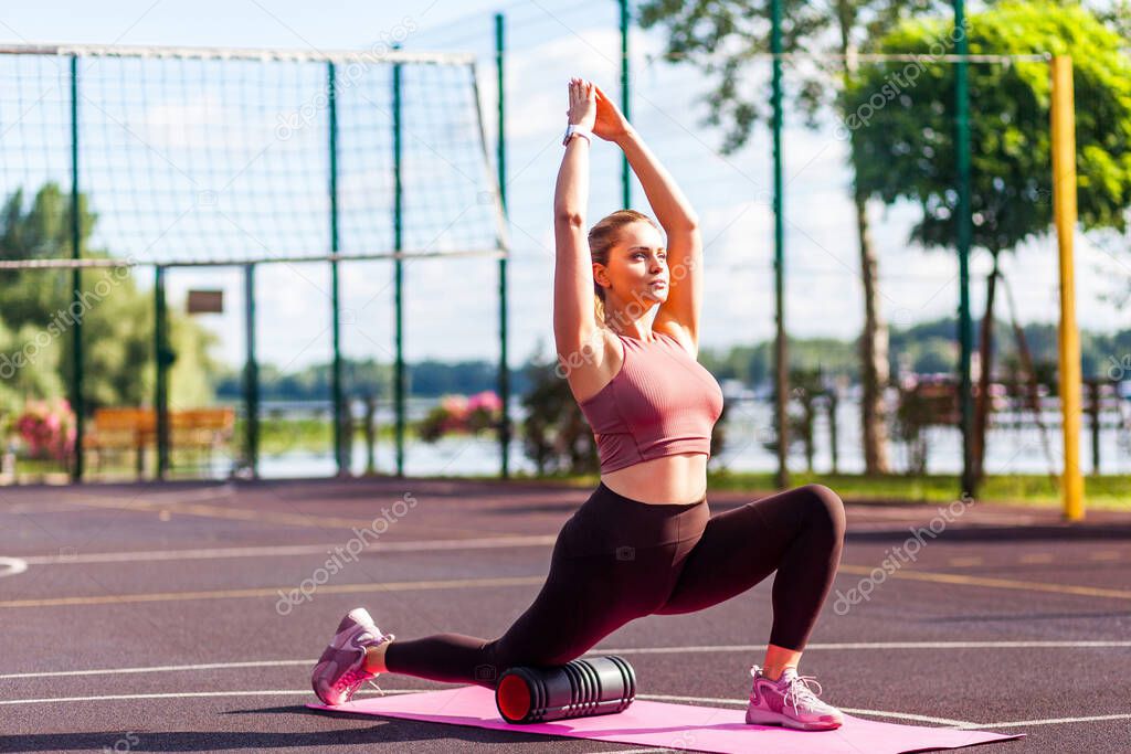 Sporty blond girl training on mat outdoor summer day, using foam roller massager to support knee while stretching hands and spinal muscles, doing fascia exercise. Health care, workouts routine