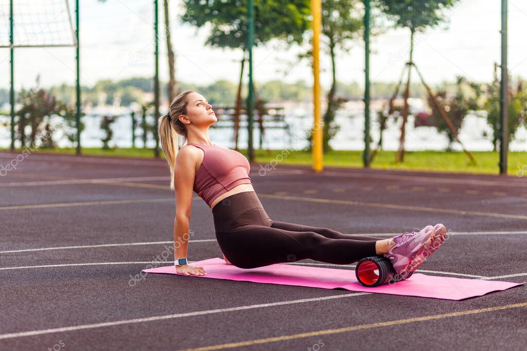 Athletic blond sporty woman training on mat outdoor summer day, massaging and stretching hamstring and ankle leg muscles on foam roller, performing fascia exercise. Health care, workouts routine