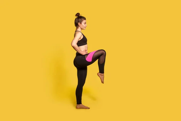 身材苗条 发髻紧的女运动员身穿紧身运动服 单腿挺立运动 大腿锻炼 锻炼肌肉 全长演播室拍摄 孤立在黄色 — 图库照片