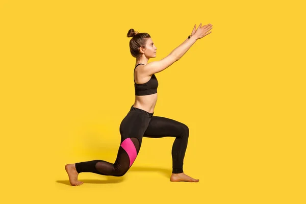 サイドビューでは タイトなスポーツウェアの中で髪パンとスリムな若い女性は 1膝立って手を上げる トレーニング筋肉を温めるスポーツラウンジ運動を行う 黄色で隔絶された長さのスタジオショット — ストック写真