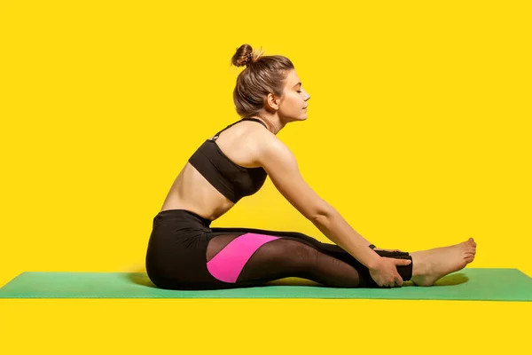 サイドビューでは タイトなスポーツウェアで髪パンを持つ女性はジムマットスポーツをやって座って 手の足の筋肉を伸ばし ウォーミングアップし 瞑想中に目を閉じておく スタジオで撮影され黄色で隔離され — ストック写真