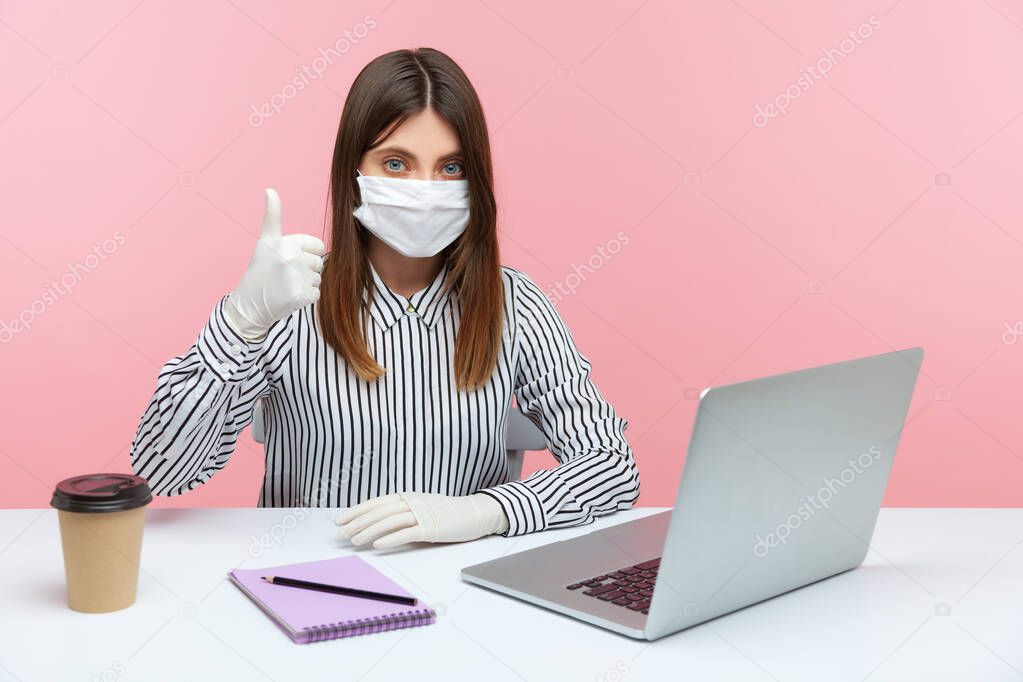Satisfied businesswoman sitting safe healthy with hygienic face mask and protective gloves during quarantine, showing thumbs up. Working at home office in self-isolation, coronavirus outbreak. indoor