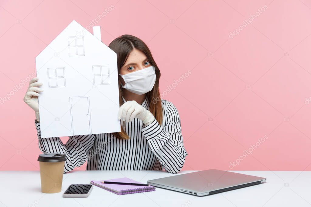 Recommendation to work home, stay safe. Woman office employee sitting at workplace, wearing hygienic mask and protective gloves, holding paper house, remote job during coronavirus quarantine. indoor