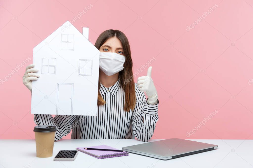Stay safe, work at home. Woman employee sitting at workplace, wearing hygienic mask and protective gloves, holding paper house and showing thumbs up, remote job during coronavirus quarantine. indoor