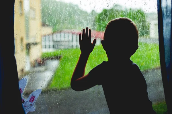 Picture of a sad young boy sitting alone at a window with hand on the glass during a quarantine.