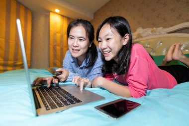 Mutlu anne ve kızı dizüstü bilgisayarlarını kullanarak evdeki yatak odasında internetten alışveriş yapmayı, not defteri ve serf web sitelerinin önünde gülümseyen Asyalı kadın ve çocuk