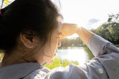 Asian woman have conjunctivitis,cataract,young female covering face by hand of bright sun in outdoor on sunny day,feel dizzy,risk of eye damage from ultraviole(UV rays),photophobia,eye health concept clipart
