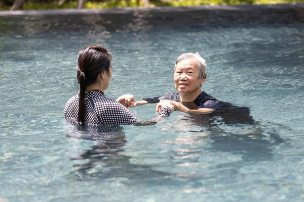 Asian caregiver woman is support,care the elderly people walking,exercising in the water,happy and healthy senior mother in swimming pool with daughter in sunny day,healthcare,physical therapy concept — Stock Photo, Image