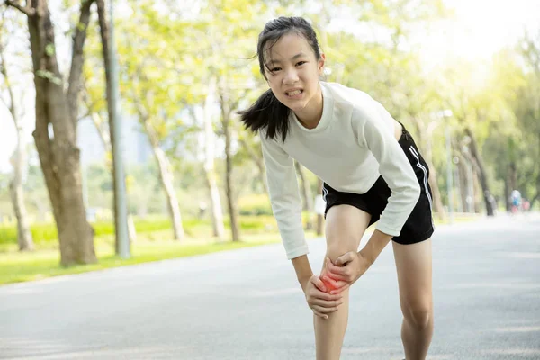 Ongelukkig aziatisch kind meisje gewond haar been pijn of kalf spier tijdens het spelen in zonnige dag, vrouwelijke tiener hand op de benen, pijn in de knie voelen van oefening in de zomer, lichamelijk letsel concept — Stockfoto