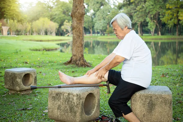 関節炎、変形性関節症、手足をマッサージするために手を使用して高齢者に苦しんで歩く棒を持つ不幸なアジアのシニア女性は、膝の痛みを感じる、自然の中で運動をした後の足の怪我 — ストック写真