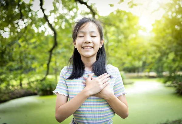 Šťastné usměvavé asijské dítě dívka stojící v zelené přírodě, držet se za ruce na srdci, cítit klid mysli, ženské dospívající zavřené oči, vychutnat dýchání čerstvého vzduchu snění, relaxovat v parku, zdravé duševní zdraví koncept — Stock fotografie