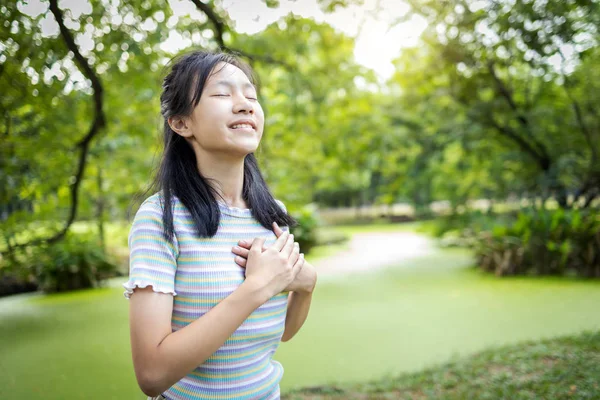 Gelukkig glimlachend aziatisch kind meisje staan in de groene natuur, vasthouden ands op hart, vrouwelijke tiener aanraken van haar borst, genieten van frisse lucht met gesloten ogen, ontspannen in het park, gezonde levensstijl concept — Stockfoto